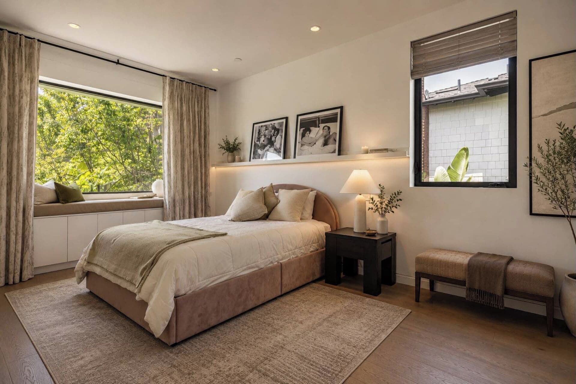 A bedroom with a bed, bedside table, bench, window, and artwork.