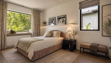 A bedroom with a bed, bedside table, bench, window, and artwork.