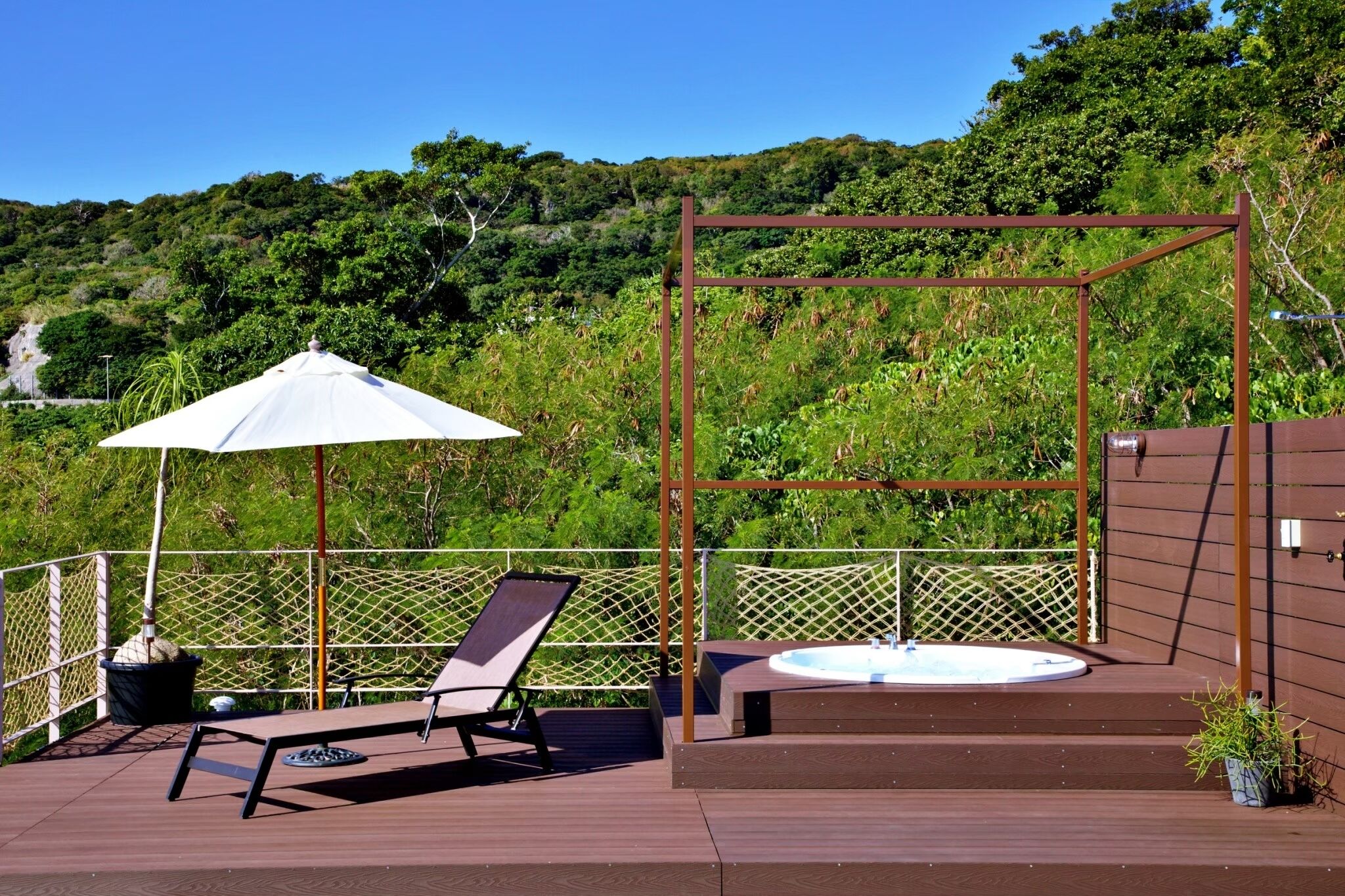 A deck with a lounge chair, a white umbrella, and a hot tub.