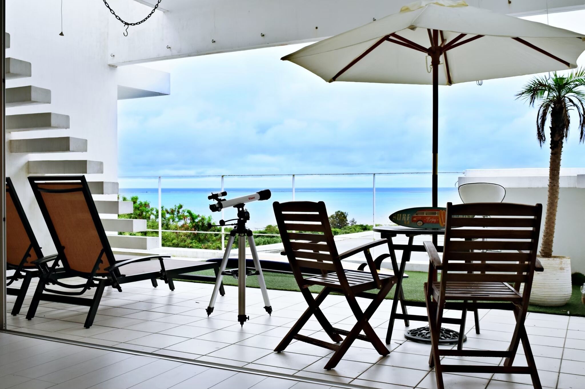 A balcony with a telescope, chairs, and a table under a large umbrella, overlooking a clear view of the sea.
