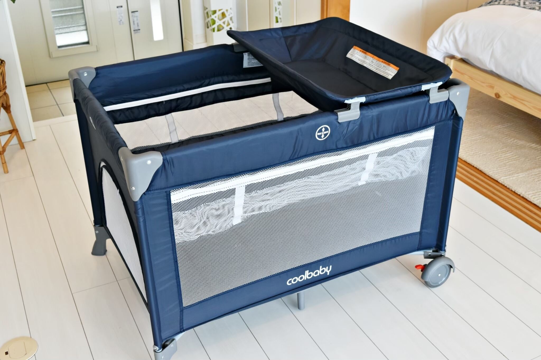 Un berceau portatif à roulettes, de la marque « coolbaby », dans une pièce où se trouvent un lit et une fenêtre.