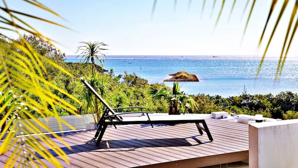 Une terrasse en bois avec un fauteuil de relaxation et un parasol en chaume, offrant une vue sur un paysage côtier avec une végétation luxuriante et la mer.