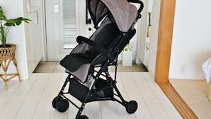 Une poussette noire pour bébé, dotée d’une capote grise, est placée sur un sol blanc, avec un tapis de sol en bois sur le côté droit.