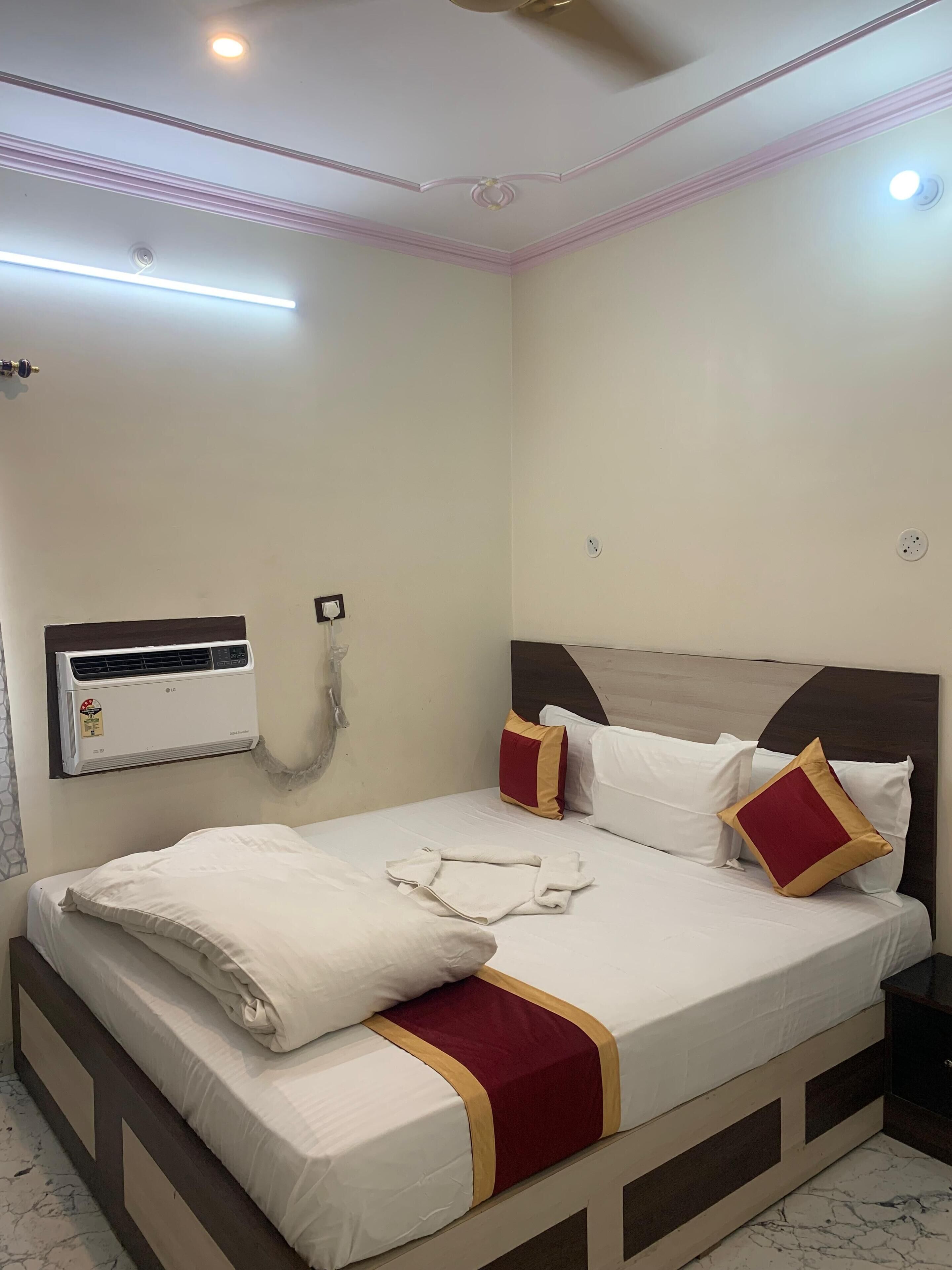A neatly made bed with white linens and a red and gold accent, a wall-mounted air conditioner, and a ceiling fan.
