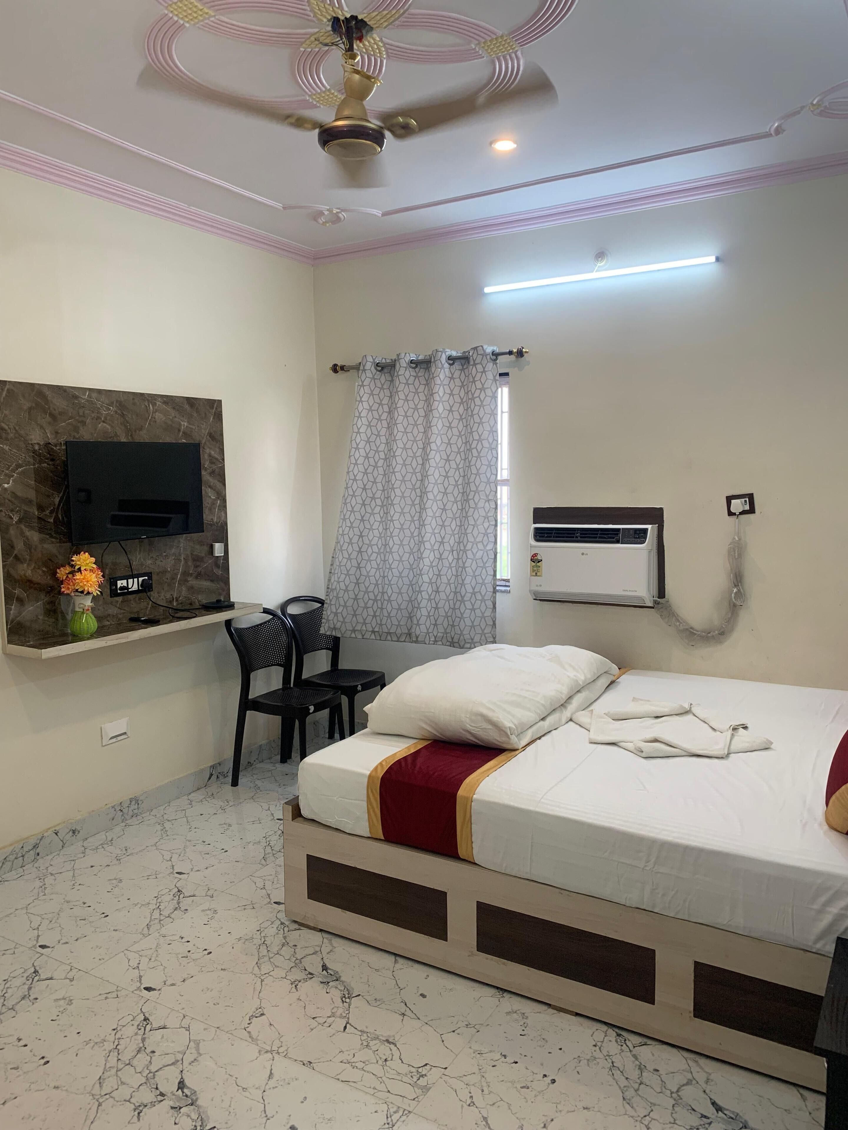 Een hotelkamer met een bed, een bureau, een stoel, een aan de muur bevestigde televisie, een plafondventilator en een airconditioning.