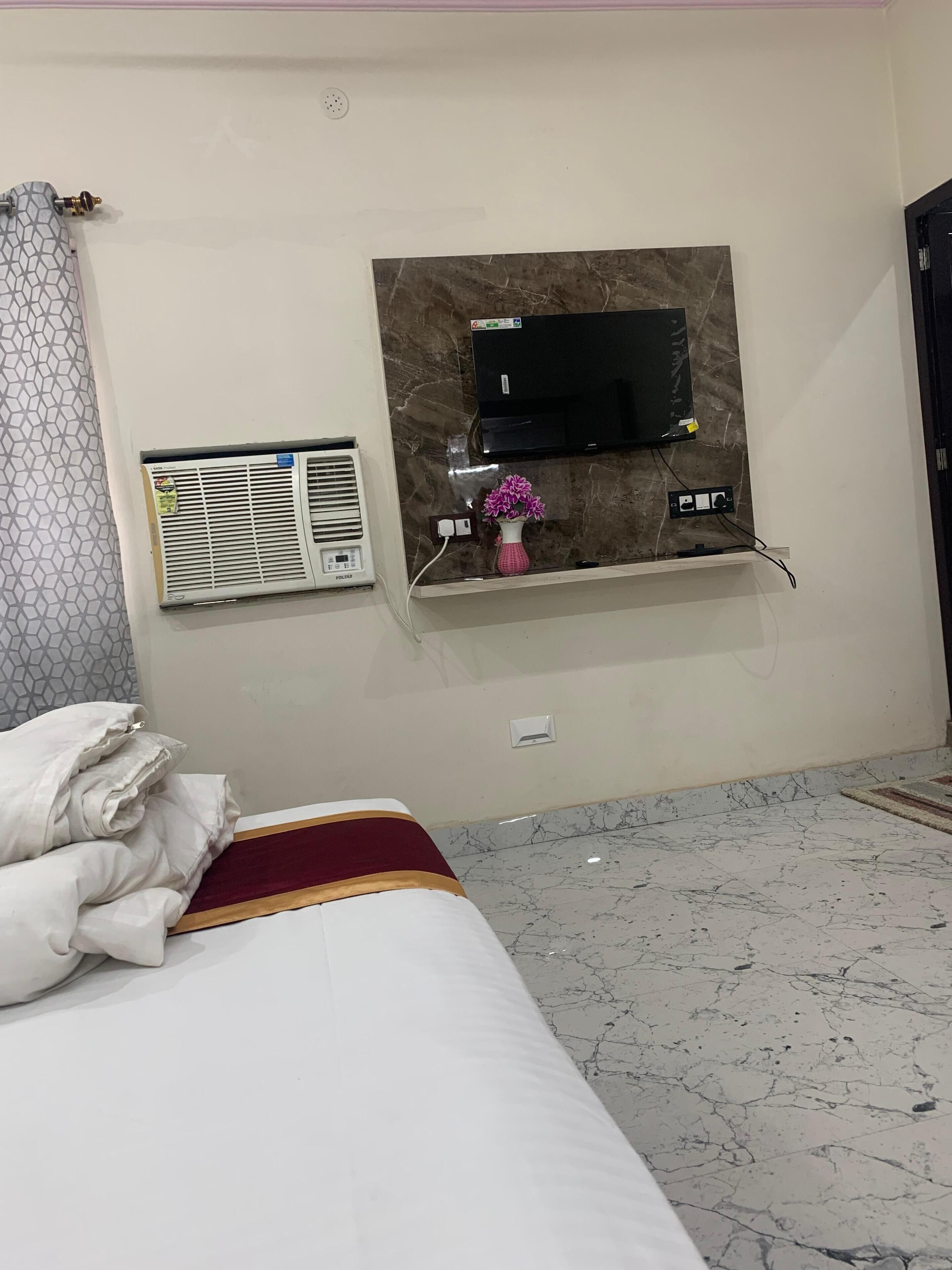 A hotel room with a bed, a wall-mounted TV, an air conditioning unit, and a power outlet.