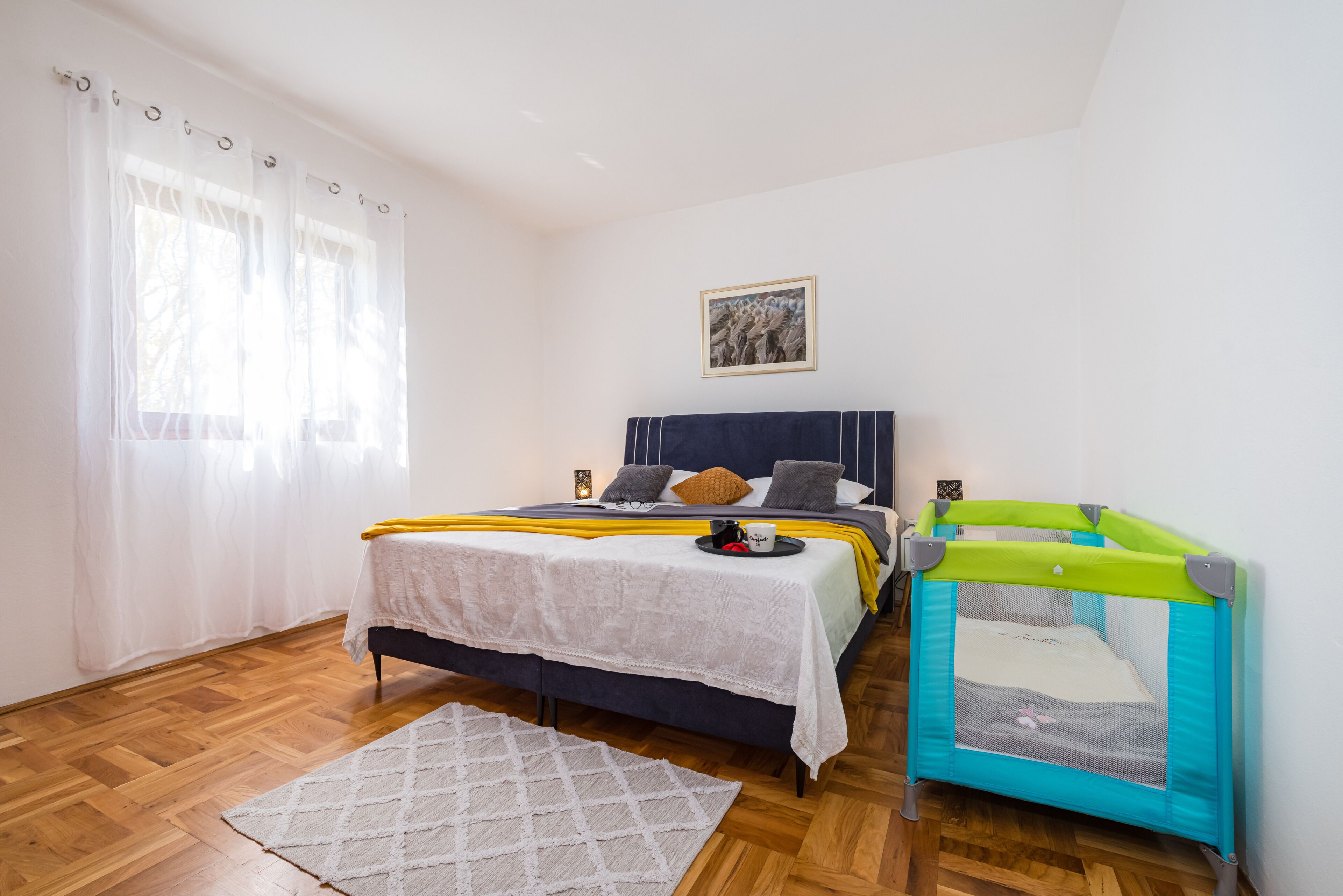Een slaapkamer met een bed, een klein ledikant, een raam met gordijnen en een schilderij aan de muur.