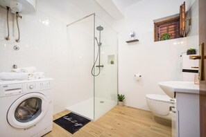 Un bany modern amb plat de dutxa envoltat de vidre, una rentadora, un vàter i una petita estant amb plantes.