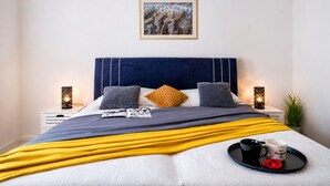 Una cama bien hecha con un cabecero azul, dos lámparas de noche, un cuadro en la pared y una bandeja con una taza y un objeto rojo sobre la cama.