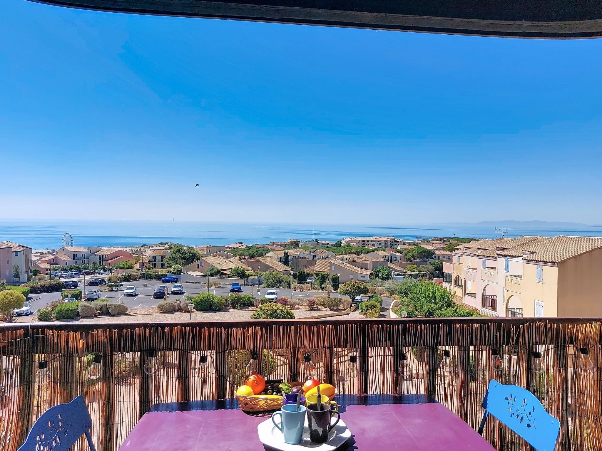 A balcony with a table set for two, overlooking a coastal town with buildings and a clear sky.