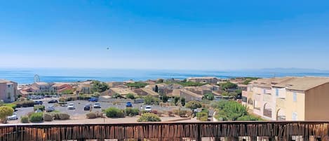 A balcony with a table set for two, overlooking a coastal town with buildings and a clear sky.