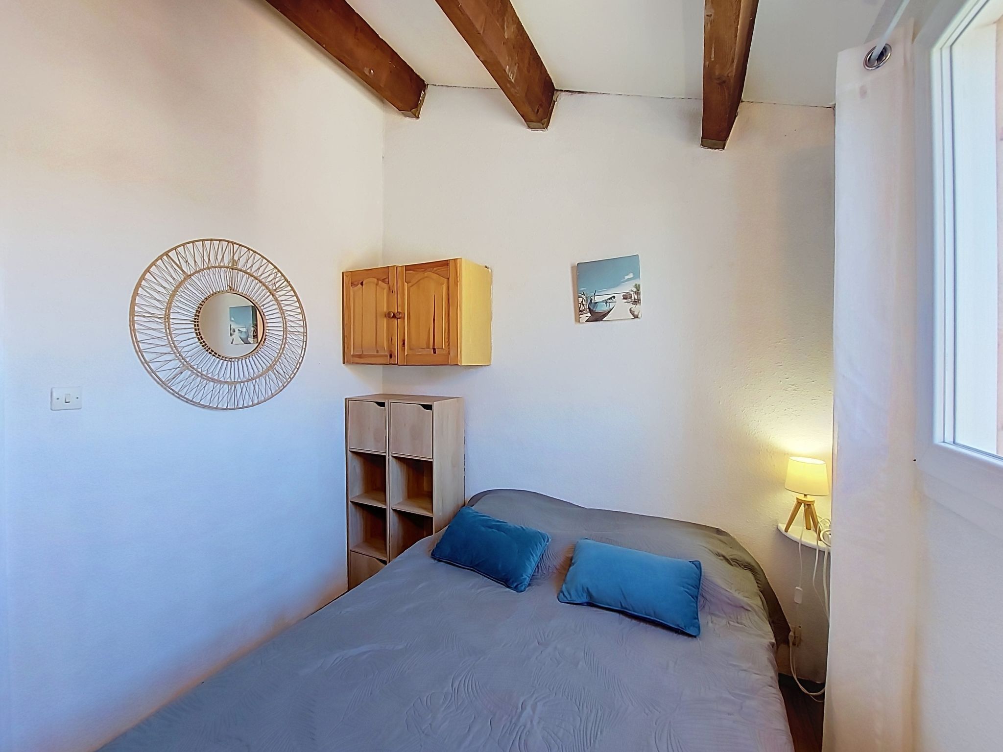 Une chambre à coucher comprenant un lit, deux oreillers, une armoire en bois, une petite étagère, un miroir fixé au mur et un tableau.