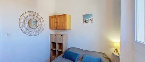 Un dormitorio con una cama, dos almohadas, un armario de madera, una pequeña estantería, un espejo en la pared y un cuadro.