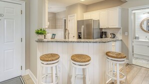Cocina con alacenas blancas, un refrigerador de acero inoxidable, encimera de granito y tres taburetes de mimbre.