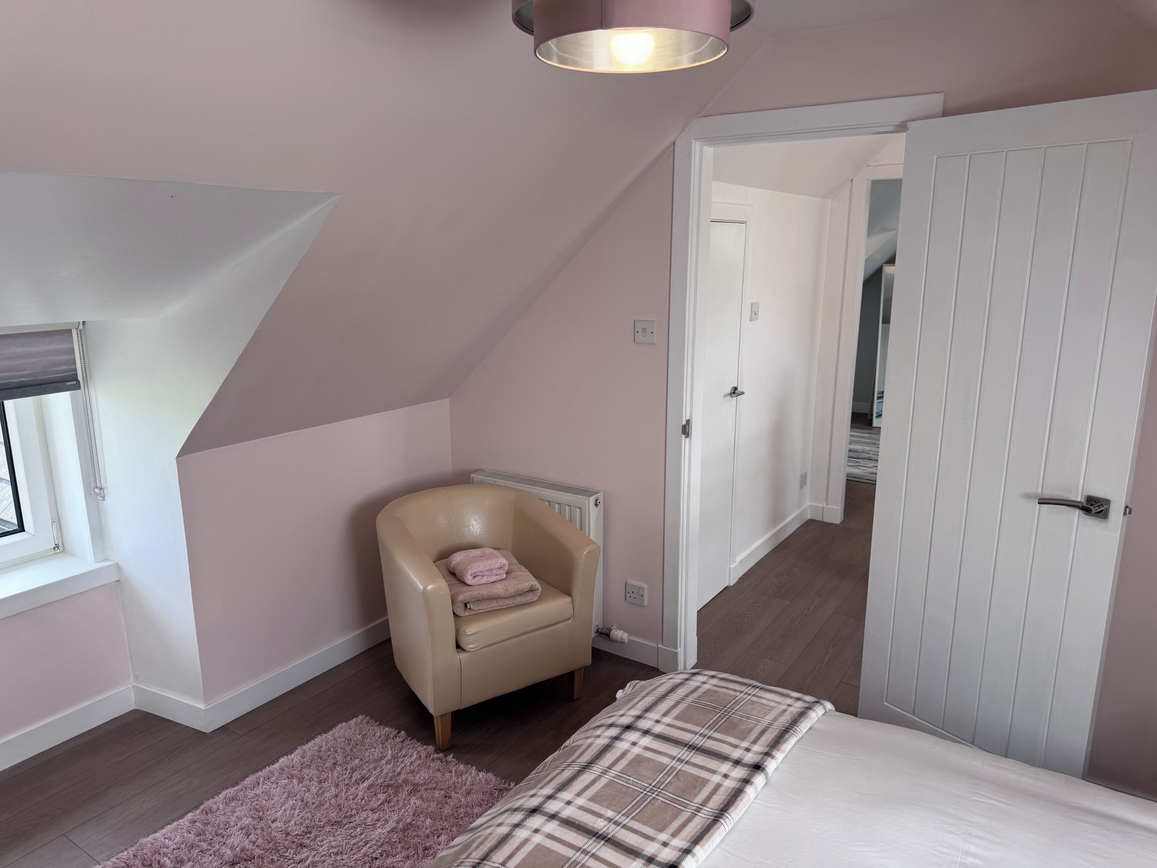 Une chambre avec un plafond en pente, une chaise recouverte d’une couverture, un lit avec un plaid, et une porte blanche.