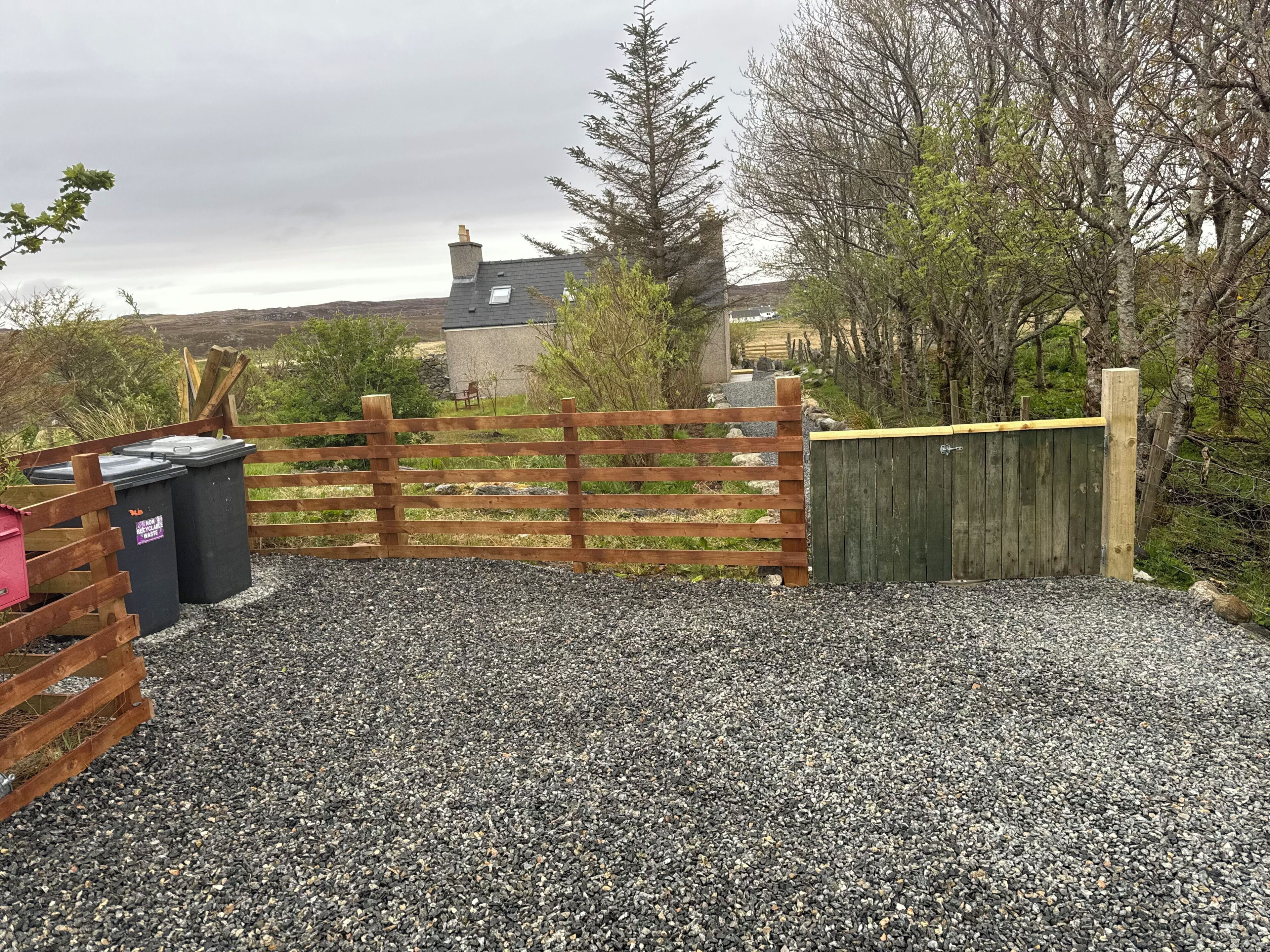 A gravel driveway leading to a wooden gate, with a stone building in the background.