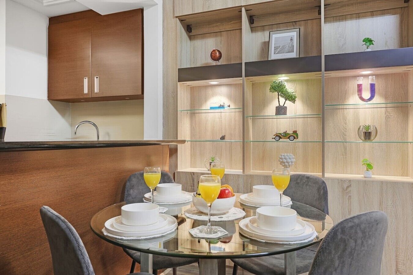 Un coin repas moderne avec une table en verre dressée pour le petit-déjeuner, des meubles en bois et des objets décoratifs disposés sur des étagères.