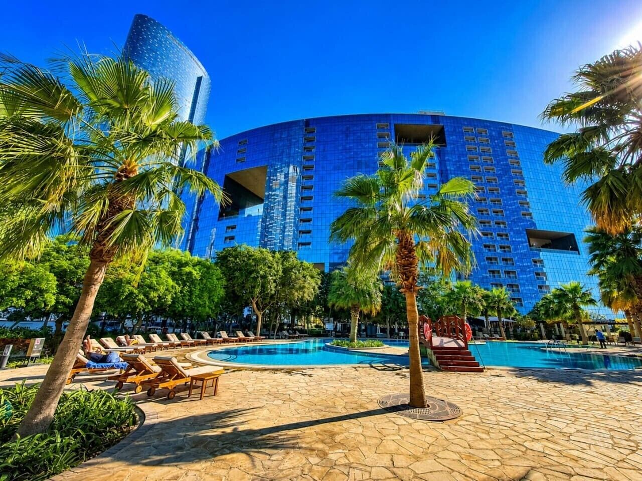 A hotel with a large pool, palm trees, and a modern blue building in the background.