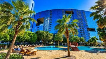 A hotel with a large pool, palm trees, and a modern blue building in the background.