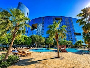 Een hotel met een groot zwembad, palmbomen en een modern blauw gebouw op de achtergrond.