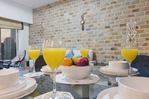 Een ontbijtsetting met fruit, sapglazen en wit servies op een glazen tafel voor een bakstenen muur.
