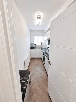 A narrow kitchen with white cabinets, a black countertop, and a herringbone wood floor. - Stylish Renovated 2BR Apartment in South London (London)
