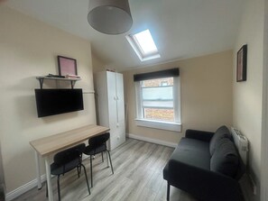 A compact living space with a desk, two chairs, a sofa, a TV mounted on the wall, a wardrobe, and a window with a view.