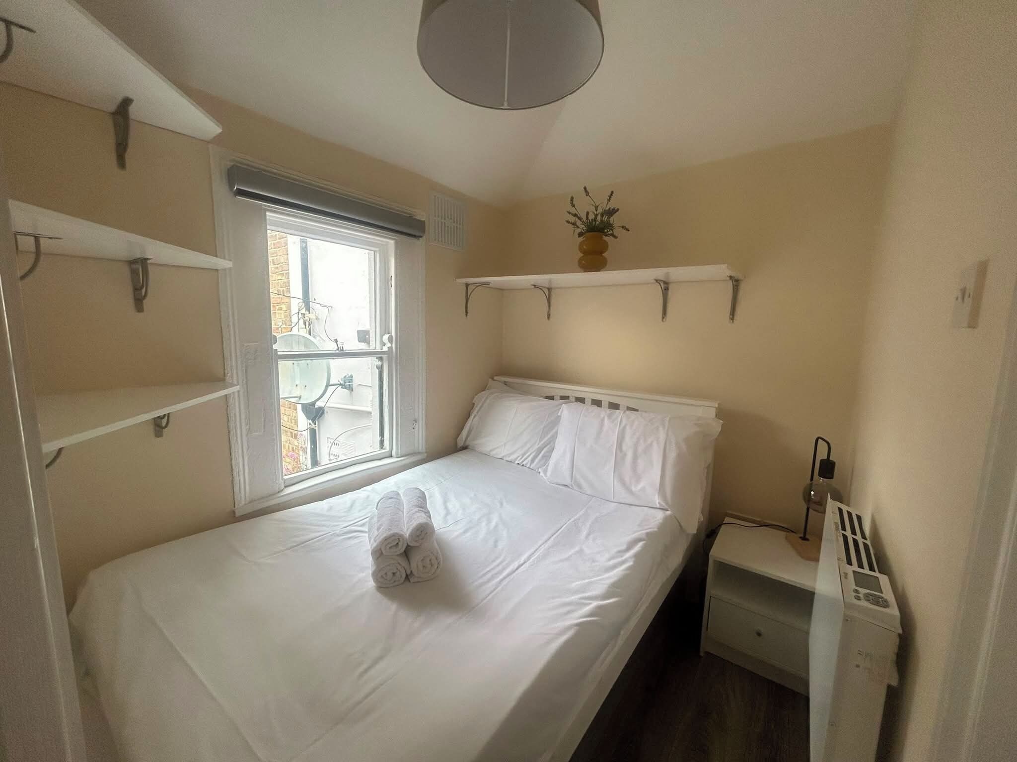 A small, neatly arranged bedroom with a single bed, a bedside table, a radiator, and a window with a view of the outside.