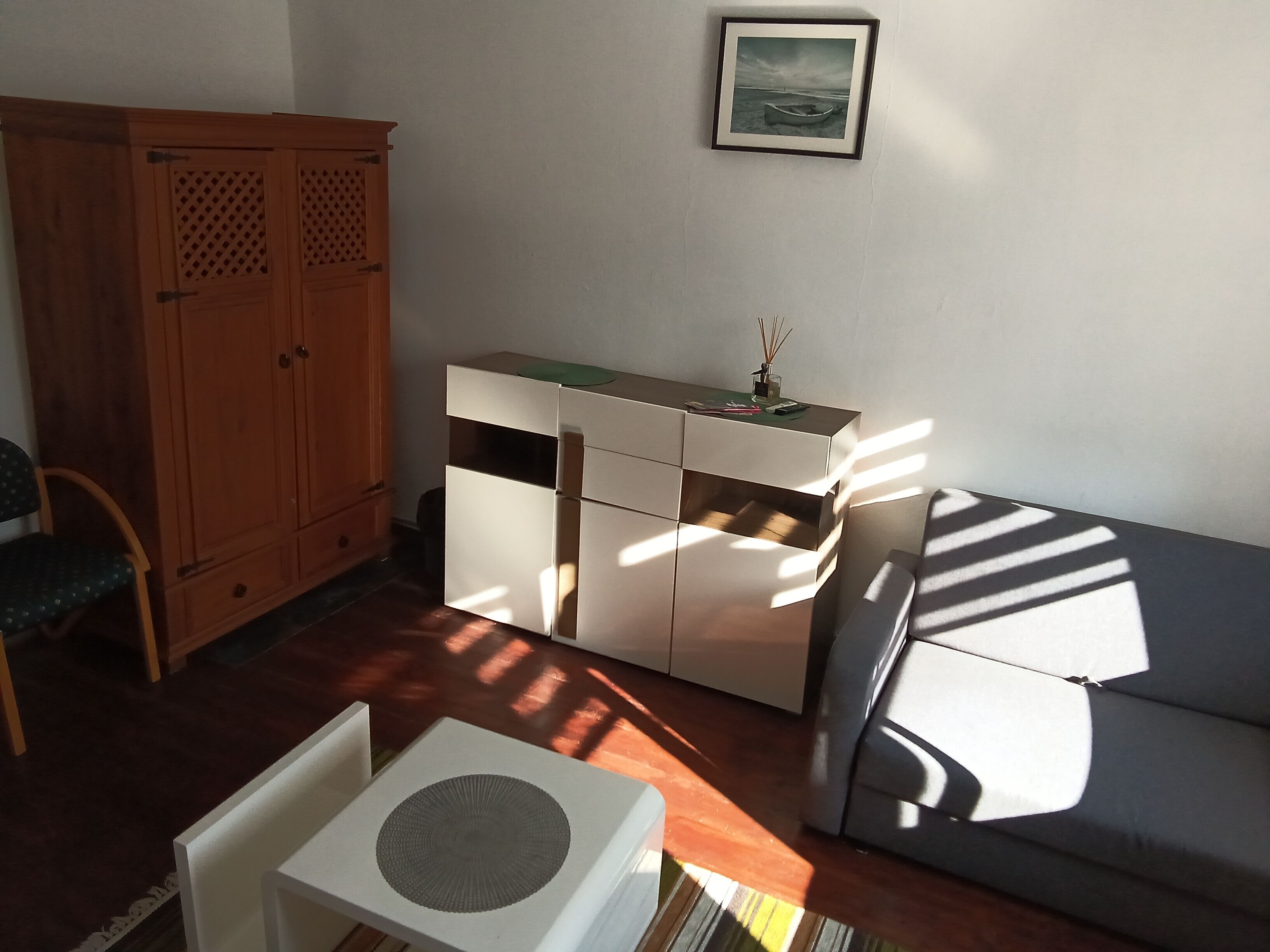 A room with a wooden wardrobe, a white cabinet, a grey sofa, and a framed picture on the wall.