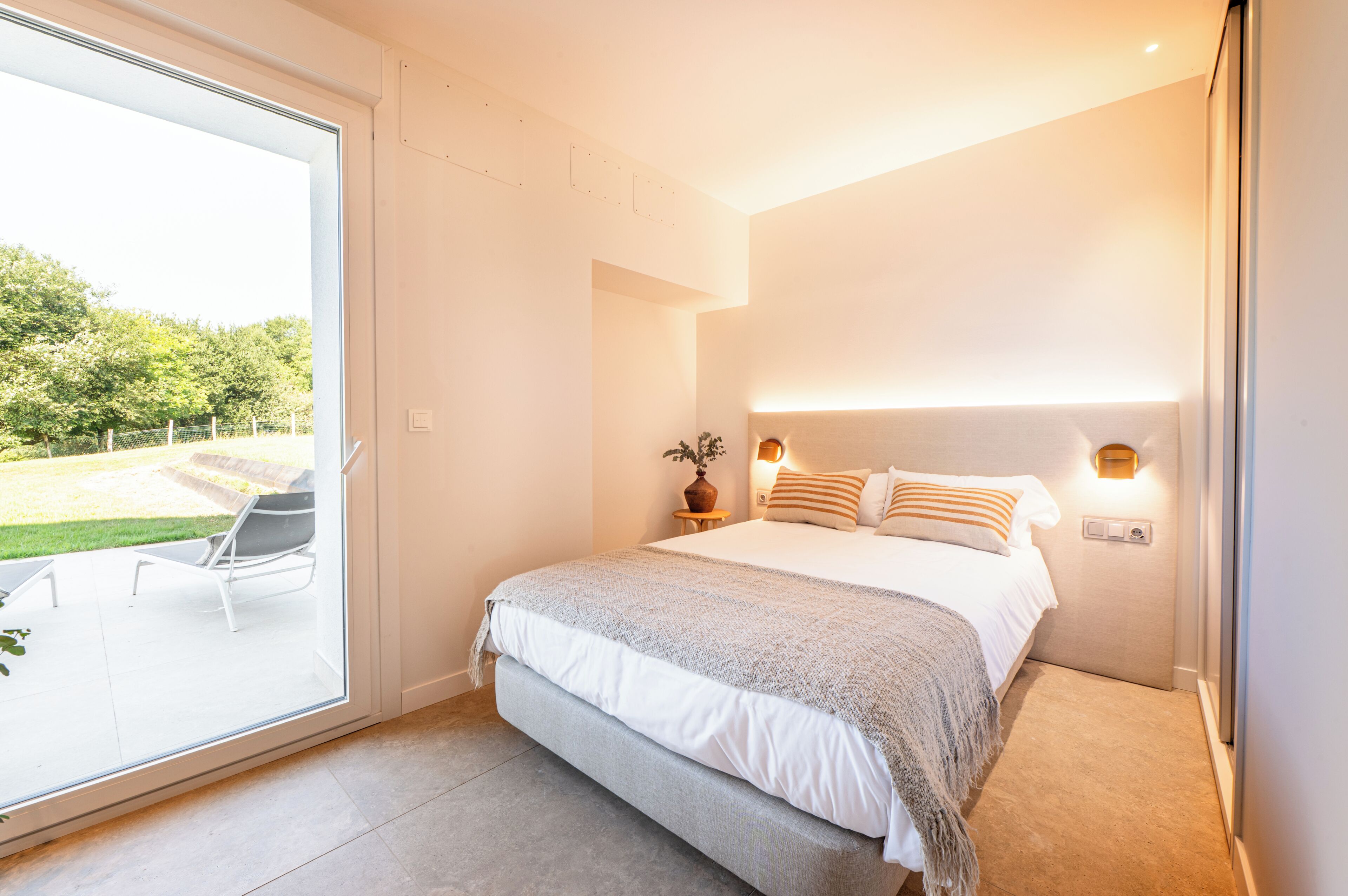 Een slaapkamer met een groot bed, een nachtkastje met een lamp en uitzicht op een tuin via een schuifdeur.