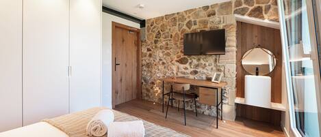 A bedroom with a bed, a desk, a chair, a TV, and a stone wall.