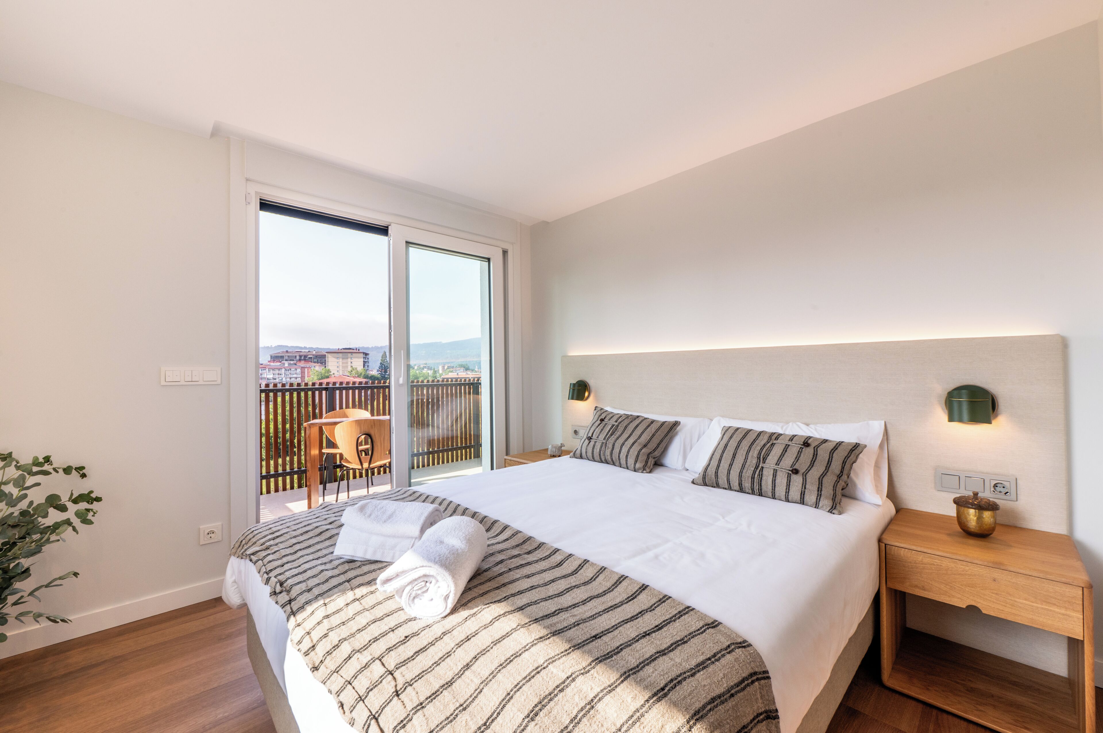 A hotel room with a large bed, a wooden nightstand, a wall-mounted lamp, and a view of a balcony with a chair and table.
