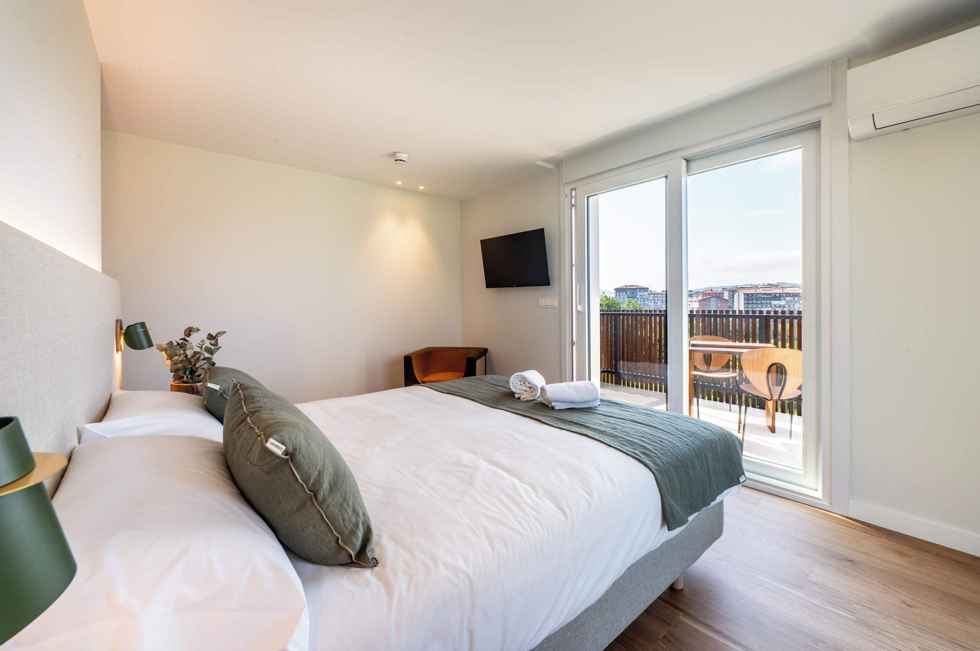 Habitación de hotel con cama, televisor, balcón con vista a edificios y piso de madera.