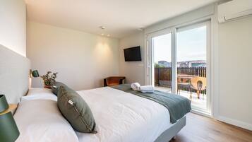 Habitación de hotel con cama, televisor, balcón con vista a edificios y piso de madera.