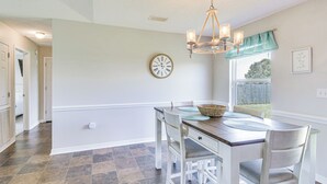 Un coin repas avec une table en bois, des chaises blanches, un lustre, une horloge et une fenêtre donnant sur un beau paysage.