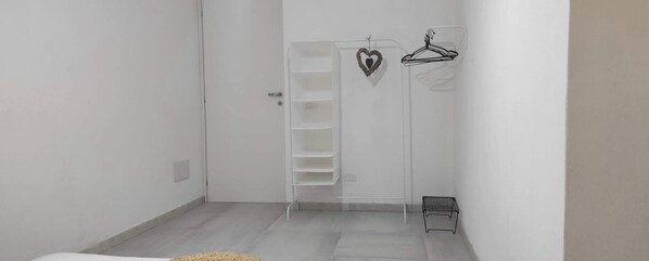 A hallway with a white door, a white closet with a hanging rod, and a small shelf unit. - The courtyard by the seaside (Calasetta)