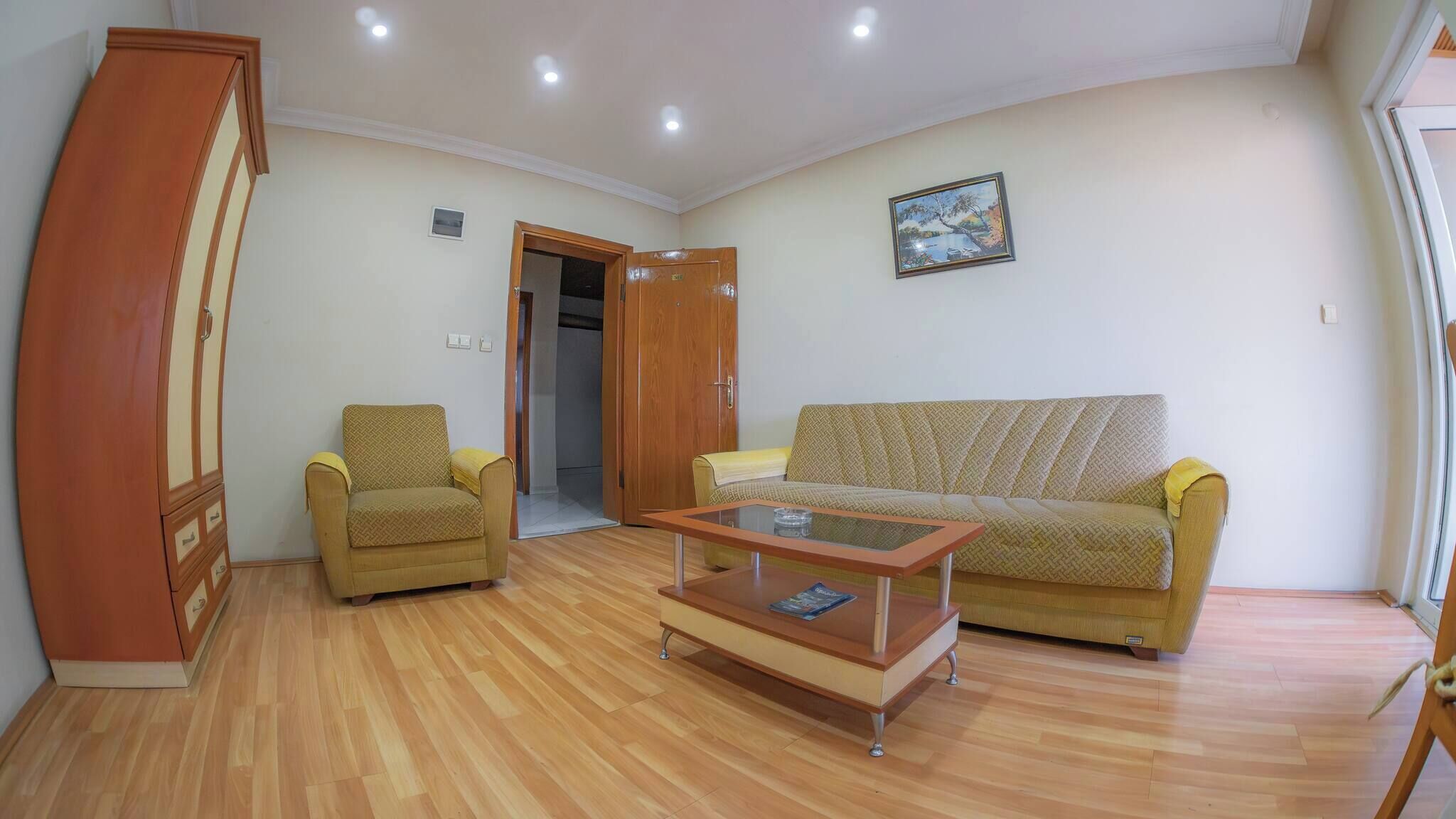 A living room with a sofa, armchair, and coffee table. There is a wooden wardrobe and a door leading to another room.