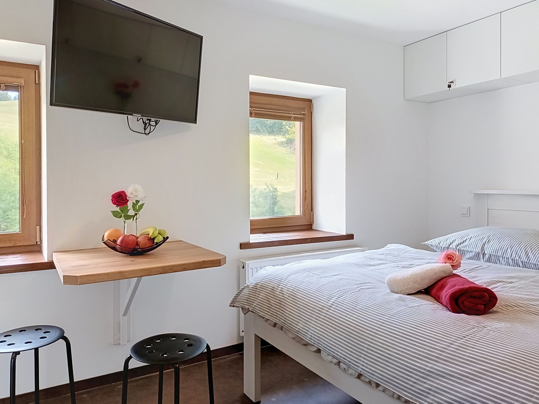 A bedroom with a bed, a TV, a window, a small table with fruit and flowers, and a chair.