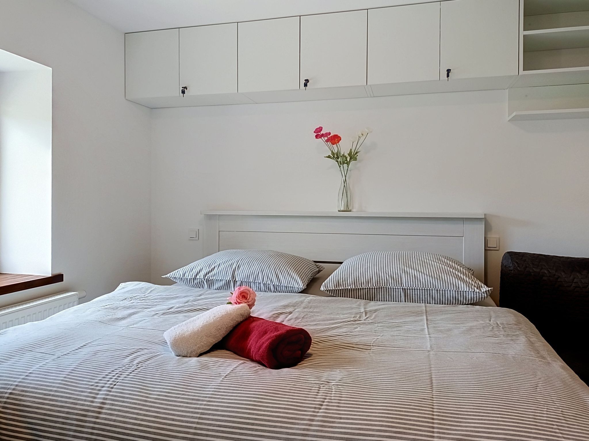 A neatly made bed with striped bedding, a vase with flowers, and a red towel on the bed.