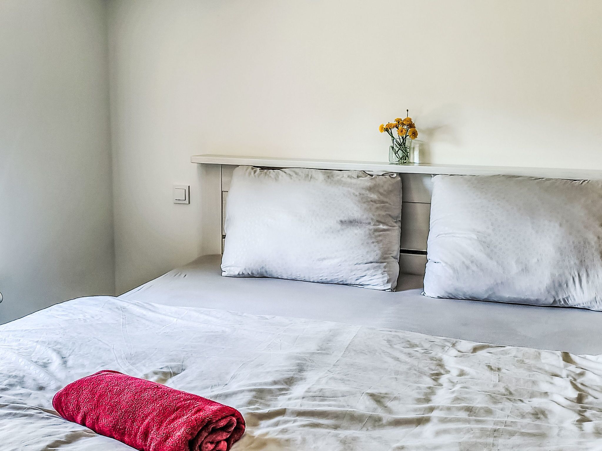 A bedroom with a bed, pillows, a bedside table with a vase of flowers, and a red towel.