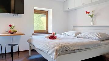 A bedroom with a bed, a desk, a chair, a TV mounted on the wall, and a window with a view of greenery.