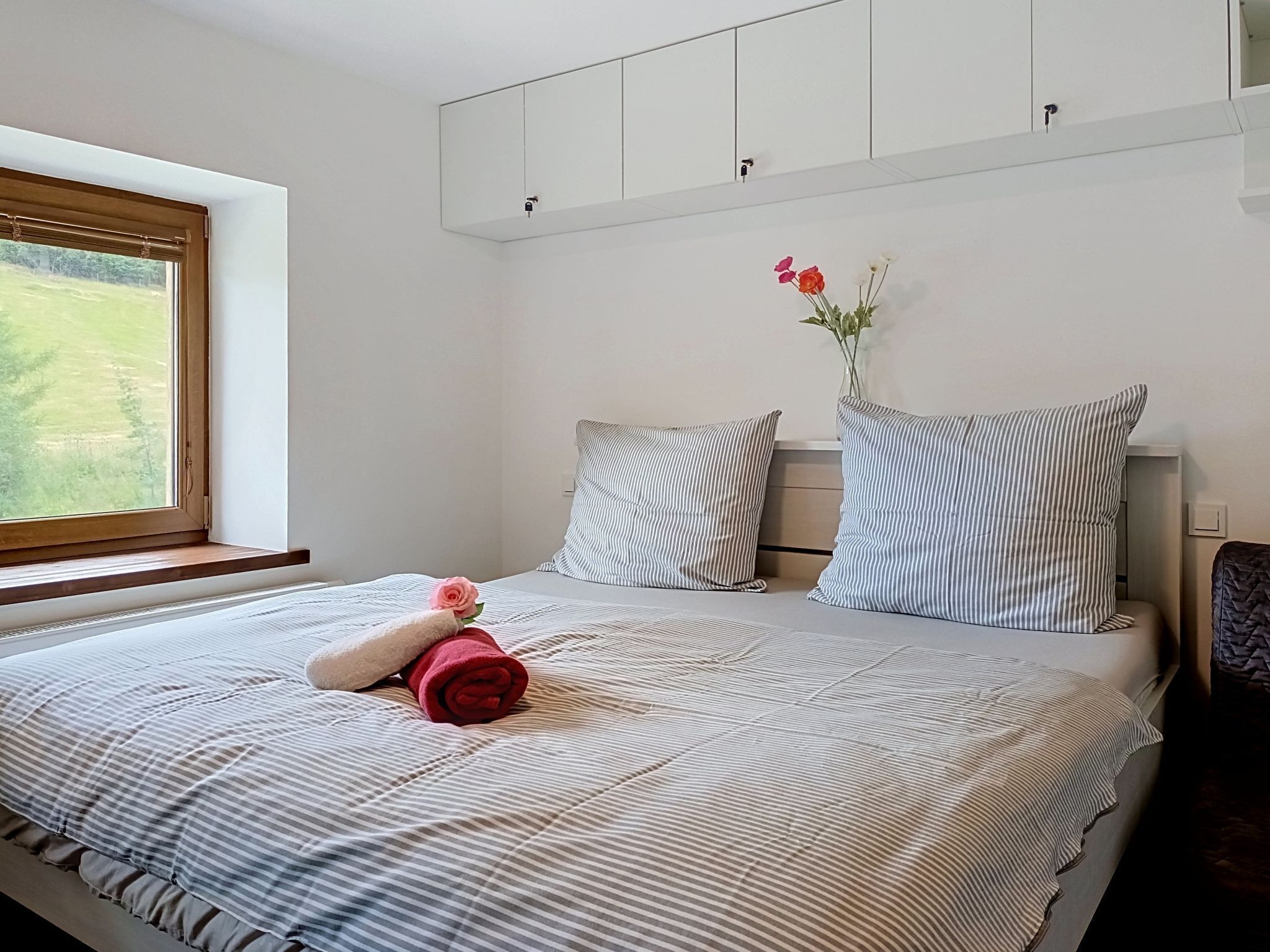 A neatly made bed with striped bedding, a window with a view of greenery, and a vase with flowers on the nightstand.