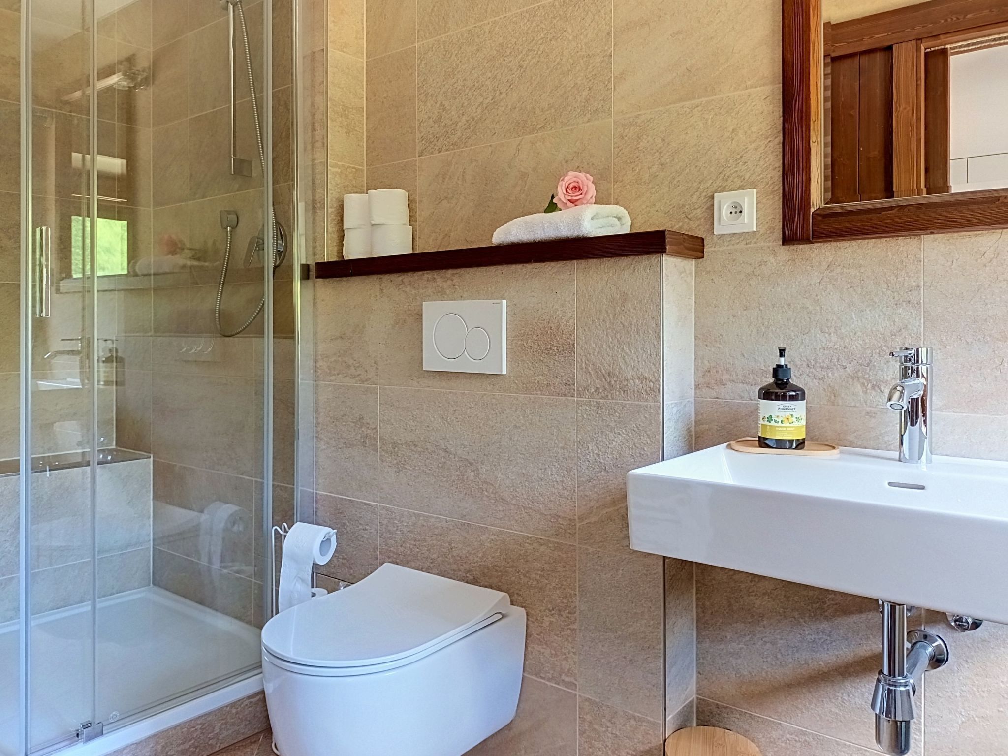 A bathroom with a glass shower enclosure, a toilet, a sink with a soap dispenser, and a towel rack with a rolled towel and a pink rose.