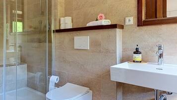 A bathroom with a glass shower enclosure, a toilet, a sink with a soap dispenser, and a towel rack with a rolled towel and a pink rose.