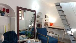 Un salon avec un fauteuil bleu, un canapé blanc, un chien, un escalier et un bureau avec une lampe.