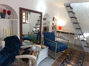 A living room with a blue armchair, a white sofa, a dog, a staircase, and a desk with a lamp. - Character Cottage with Balcony in Peaceful Garden (Johannesburg)