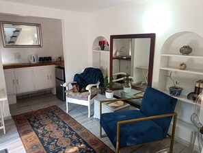 A living room with a blue armchair, a white sofa, a coffee table, and a rug. There is a kitchen area with cabinets and a refrigerator. - Character Cottage with Balcony in Peaceful Garden (Johannesburg)