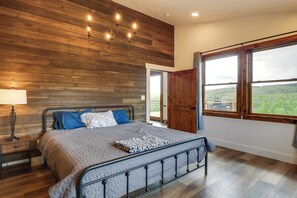 A bedroom with a wooden wall, a bed with a metal frame, a nightstand, a lamp, and a window with a view. - On 7 Acres! Scenic Home w/ Game Room in Fairplay (Fairplay)