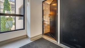 Una habitación de hotel moderna con un ventanal grande, una puerta que da a un pasillo y un balcón visible.