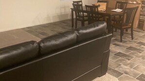 Un salon avec un canapé en cuir marron foncé, un coin repas avec des chaises en bois et une table, et une fenêtre avec des stores.